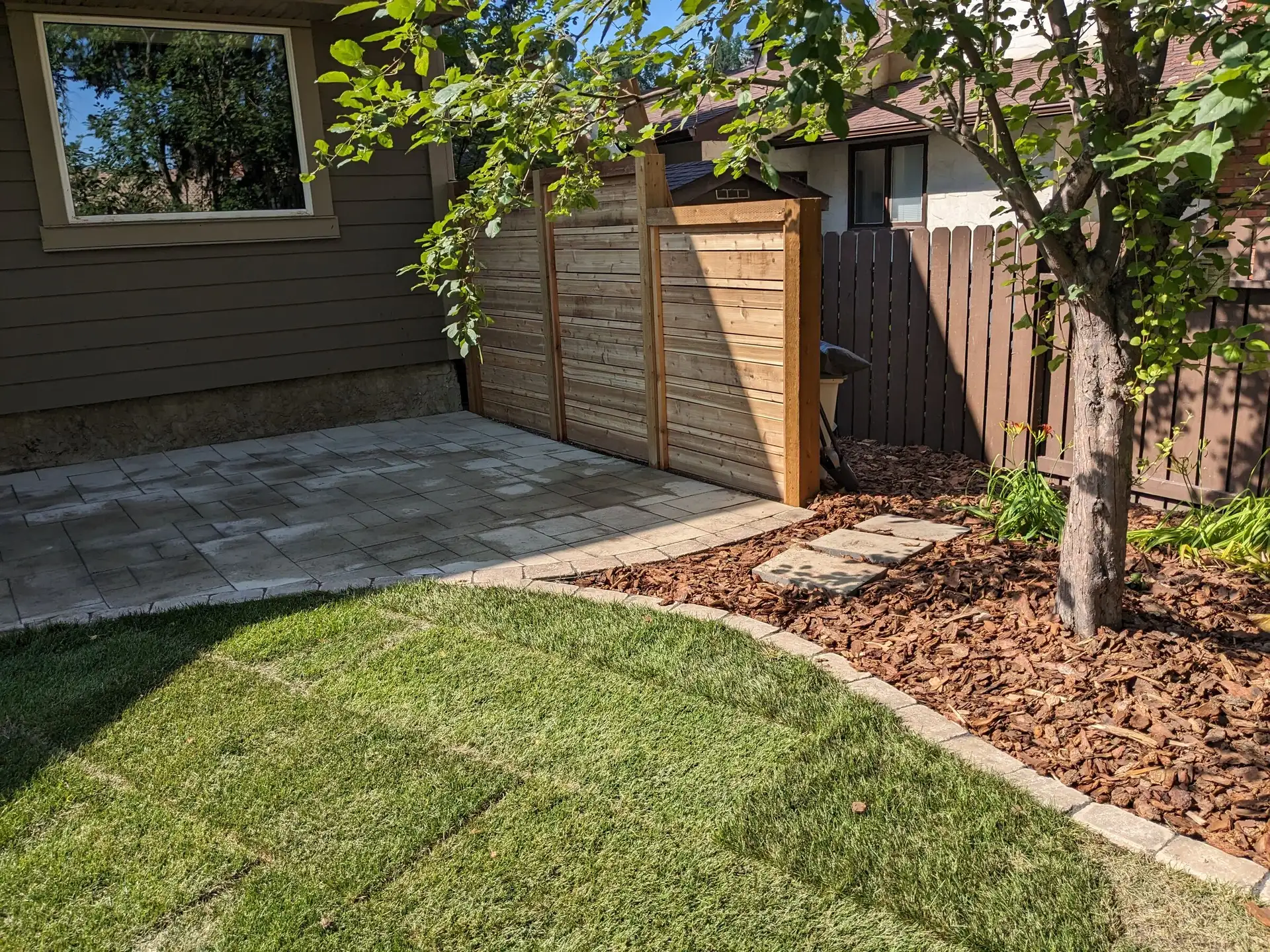 Curved garden bed with mulch and paver edging around a tree, next to new sod and a paver patio
